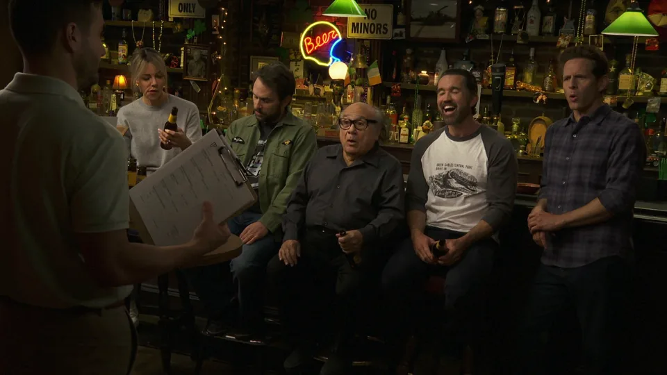 The gang sits at Paddy’s Pub with bottled beer while a sales representative holds a clipboard.